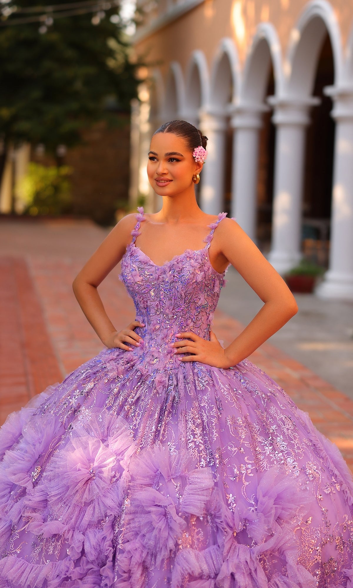 A young woman in the Quinceanera Dress 54314 By Amarra, a detailed lavender ball gown, stands outdoors on a brick path, smiling with her hands on her hips.